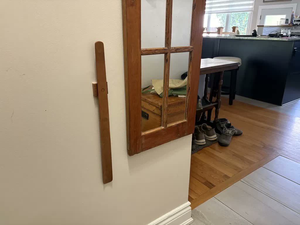 Skohorn i ljus ek på cremefärgad vägg bredvid en gammal spegel i rå träram, med utsikt in i köket