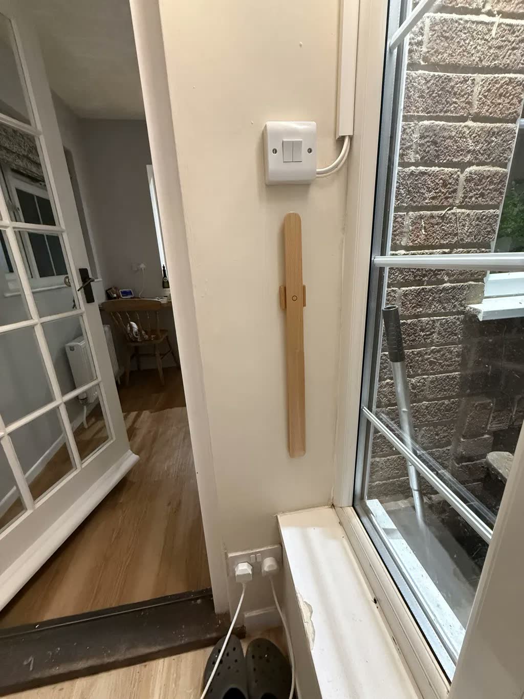 Skohorn i ljus ek på cremefärgad vägg vid fönster och dörröppning, med strömbrytare ovanför och träskor på golvet